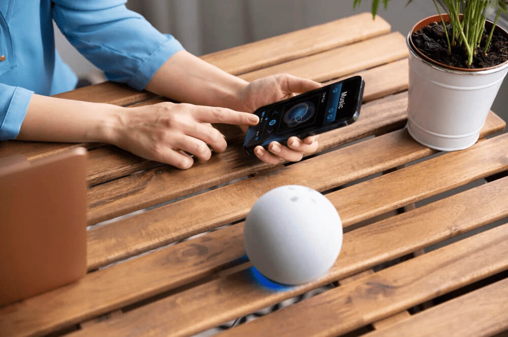 Person steuert Musik per Smartphone-App und sprachgesteuertem Smart Speaker auf einem Holztisch.