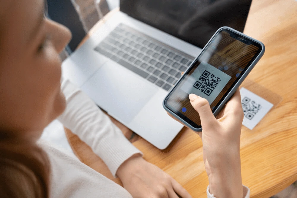 Frau scannt mit ihrem Smartphone einen QR-Code neben einem Laptop, der auf einem Holztisch steht.