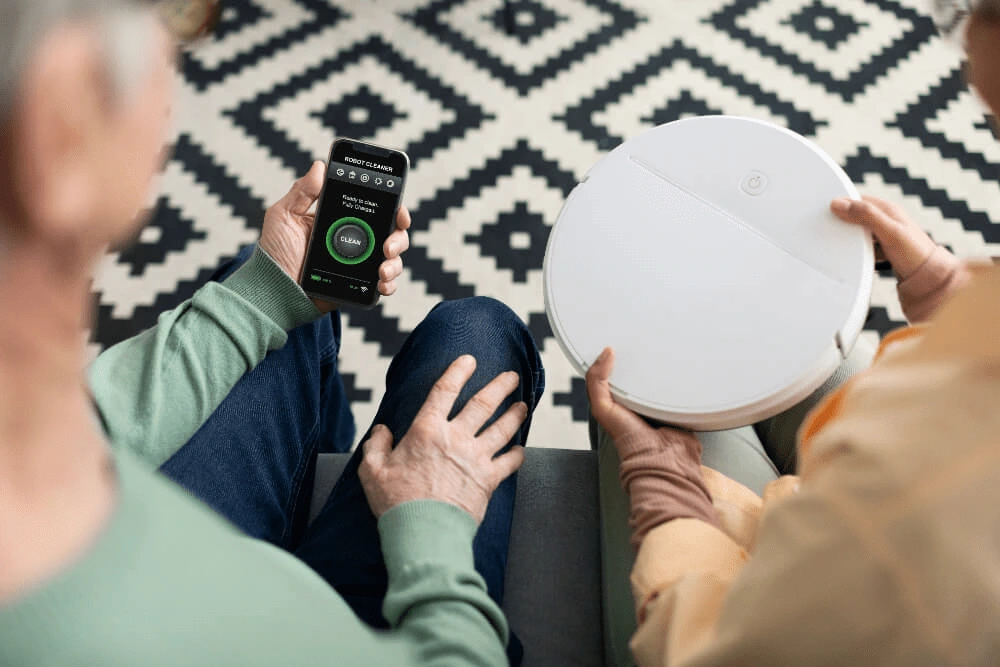 Zwei Personen sitzen auf einem Sofa, halten einen weißen Saugroboter und bedienen ihn über eine Smartphone-App.    Matter 2025