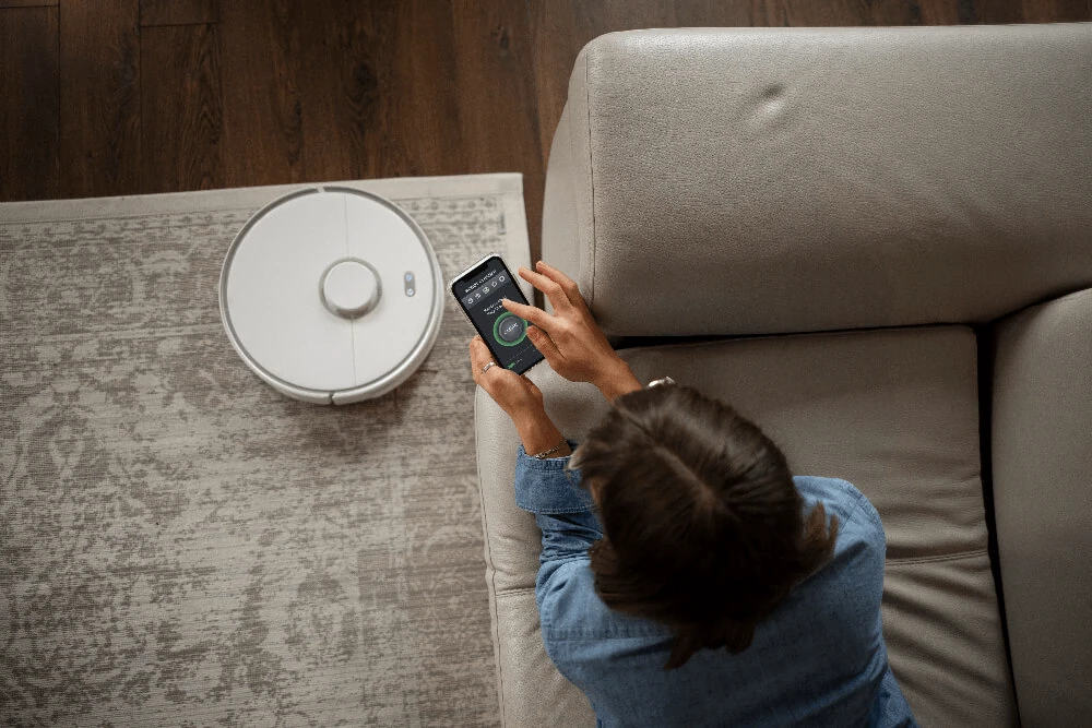 Frau sitzt auf einem Sofa und steuert einen Saugroboter per Smartphone-App in einem modernen Wohnzimmer mit Teppichboden.
