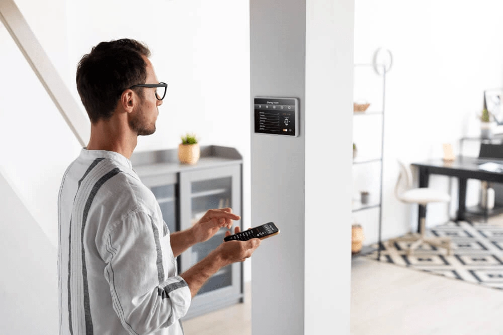 Mann bedient ein zentrales Smart-Home-Bedienfeld an der Wand mit zusätzlicher Steuerung über ein Smartphone in einem hell eingerichteten Wohnraum.